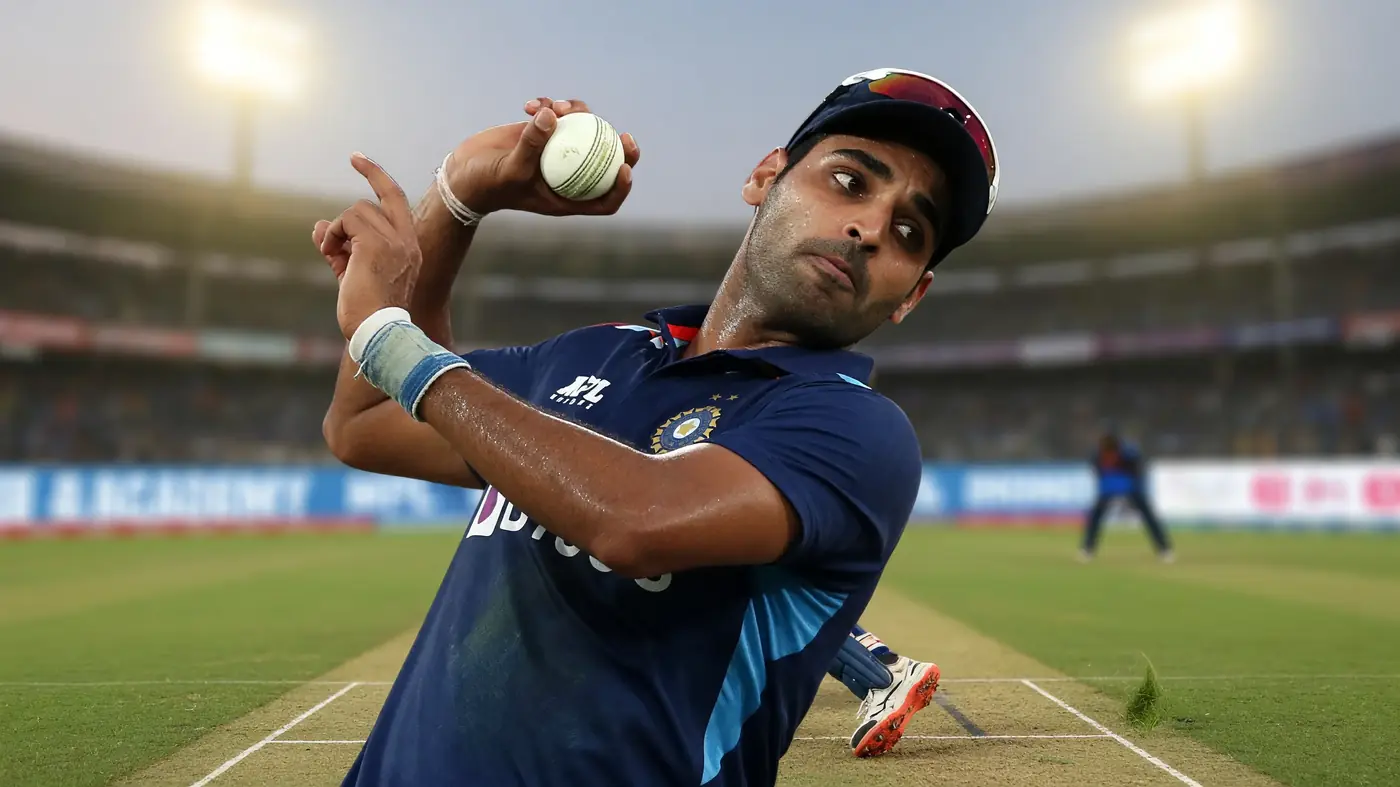 Pace bowler in a dark blue kit releasing the ball in his delivery stride under stadium floodlights